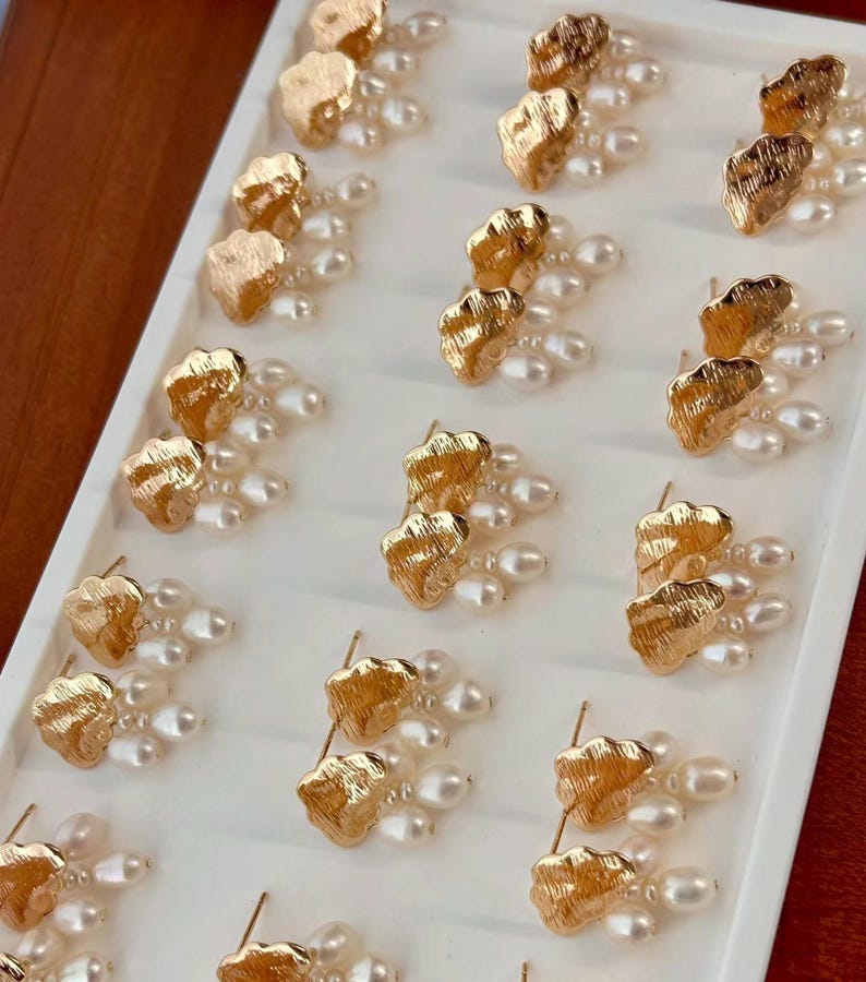 Puede incluir: Una muestra de pendientes de tono dorado con detalles de perlas. Cada pendiente presenta un dise&ntilde;o texturizado en forma de nube con tres perlas colgantes.