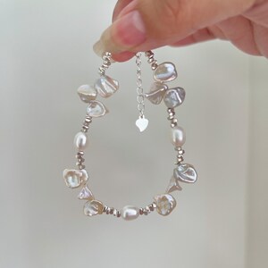 May include: A bracelet with a mix of irregular, iridescent pearls and small, silver beads. The bracelet is held up, showing its design and the adjustable chain with a heart-shaped charm. The pearls have a soft, natural lustre.