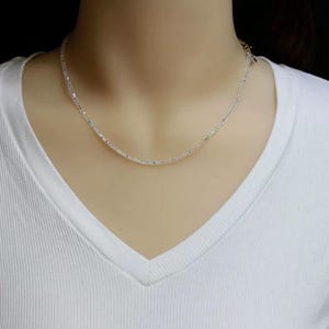 May include: A delicate silver necklace with a subtle sparkle. The necklace is composed of small, evenly spaced beads, creating a minimalist and elegant design. The necklace is displayed against a white v-neck shirt.