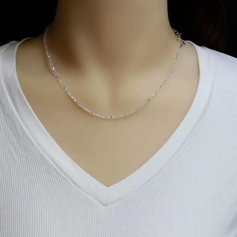 May include: A delicate silver necklace with a subtle sparkle. The necklace is composed of small, evenly spaced beads, creating a minimalist and elegant design. The necklace is displayed against a white v-neck shirt.