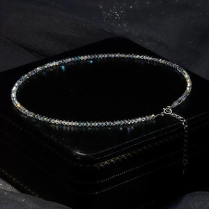 May include: A delicate necklace composed of small, iridescent beads. The necklace is displayed on a black surface, showcasing its shimmering qualities. The clasp and extender chain are silver-toned, adding a subtle contrast to the overall design.