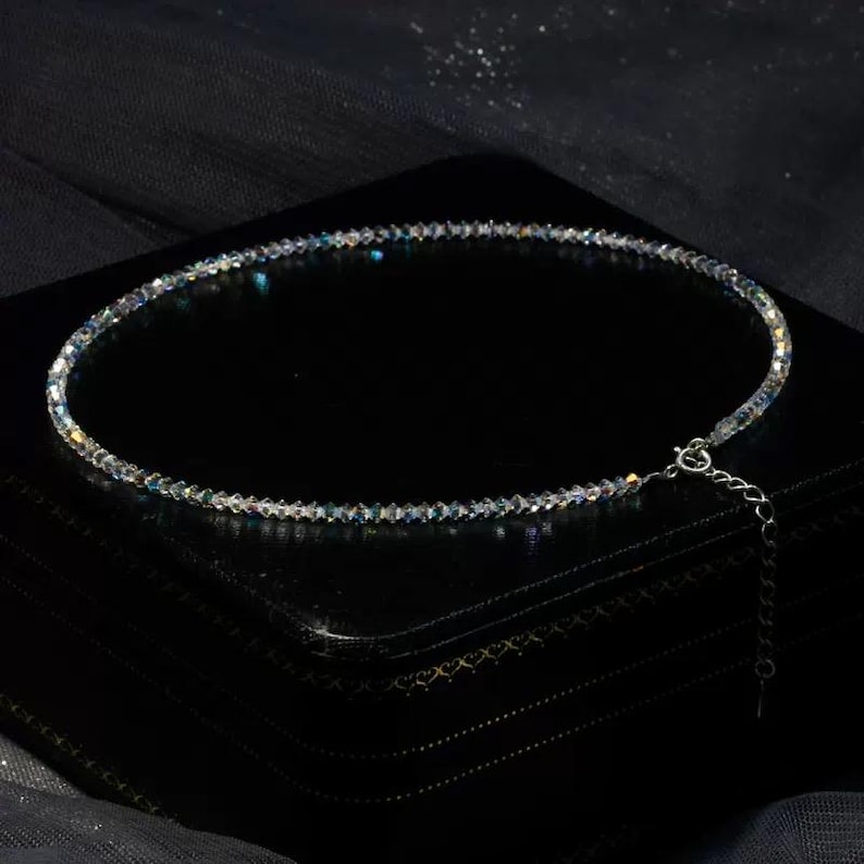May include: A delicate necklace composed of small, iridescent beads. The necklace is displayed on a black surface, showcasing its shimmering qualities. The clasp and extender chain are silver-toned, adding a subtle contrast to the overall design.