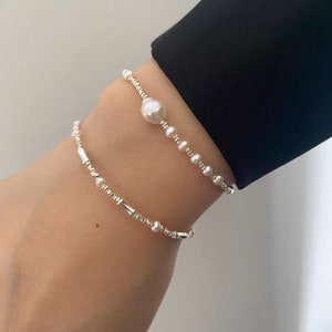 May include: Two silver bracelets with small white pearls and a larger pearl charm. The bracelets are layered on a person's wrist.