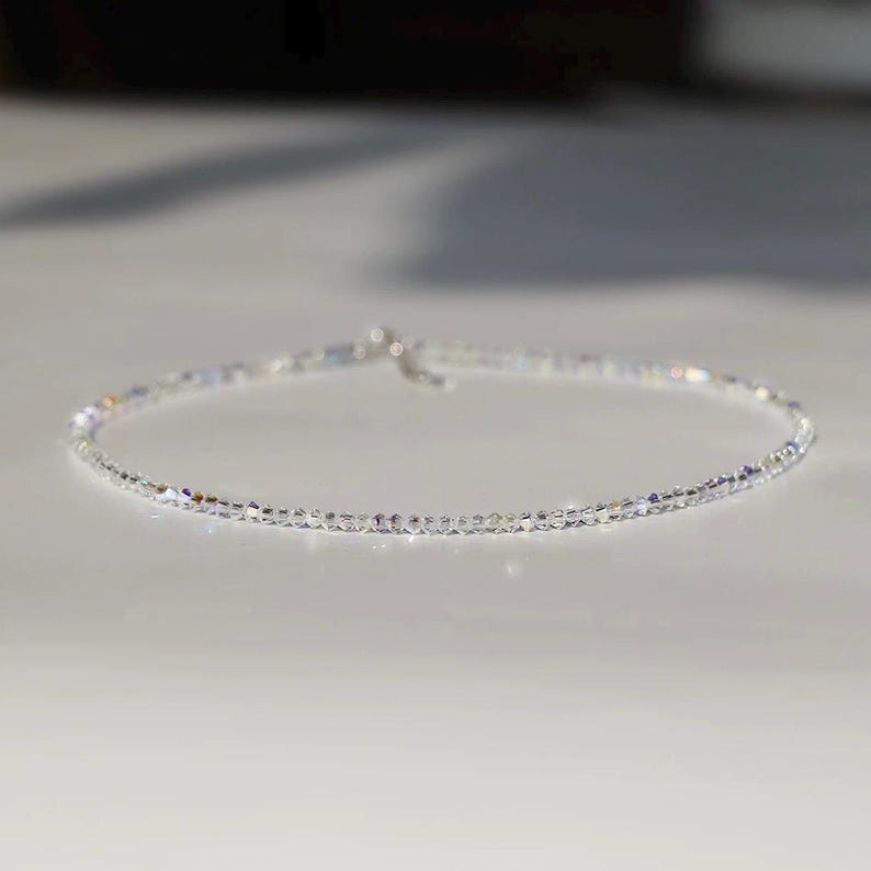 May include: A delicate, clear crystal necklace. The necklace is composed of small, sparkling crystals strung together, creating a shimmering effect. The clasp is silver. The necklace is displayed on a white surface.