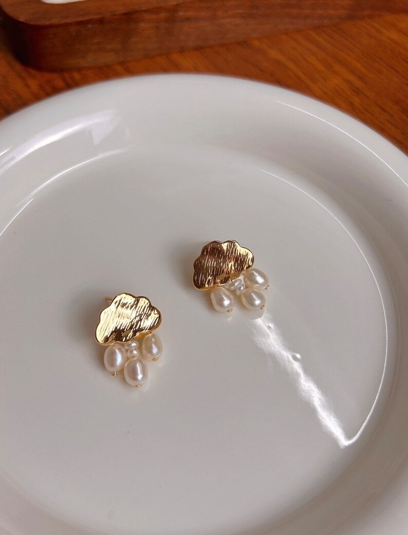 Puede incluir: Un par de pendientes de nube de oro con gotas de perlas colgantes. Los pendientes est&aacute;n en un plato blanco.