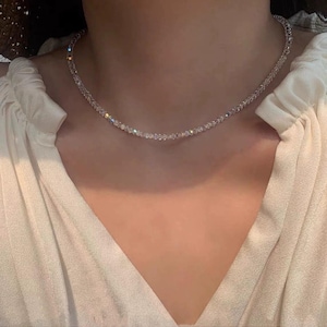 May include: A delicate, clear crystal necklace. The necklace is composed of small, faceted beads that catch the light, creating a sparkling effect. The necklace is worn against a white blouse.