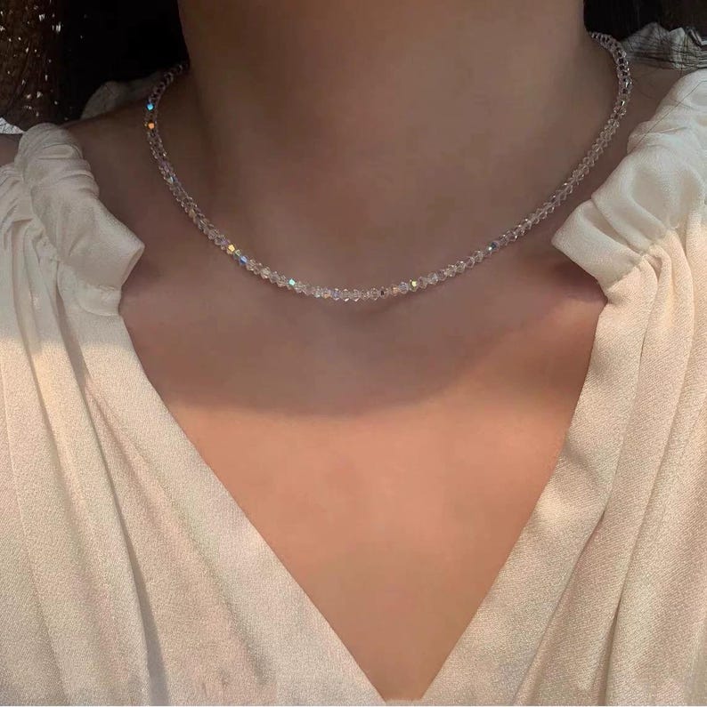 May include: A delicate, clear crystal necklace. The necklace is composed of small, faceted beads that catch the light, creating a sparkling effect. The necklace is worn against a white blouse.