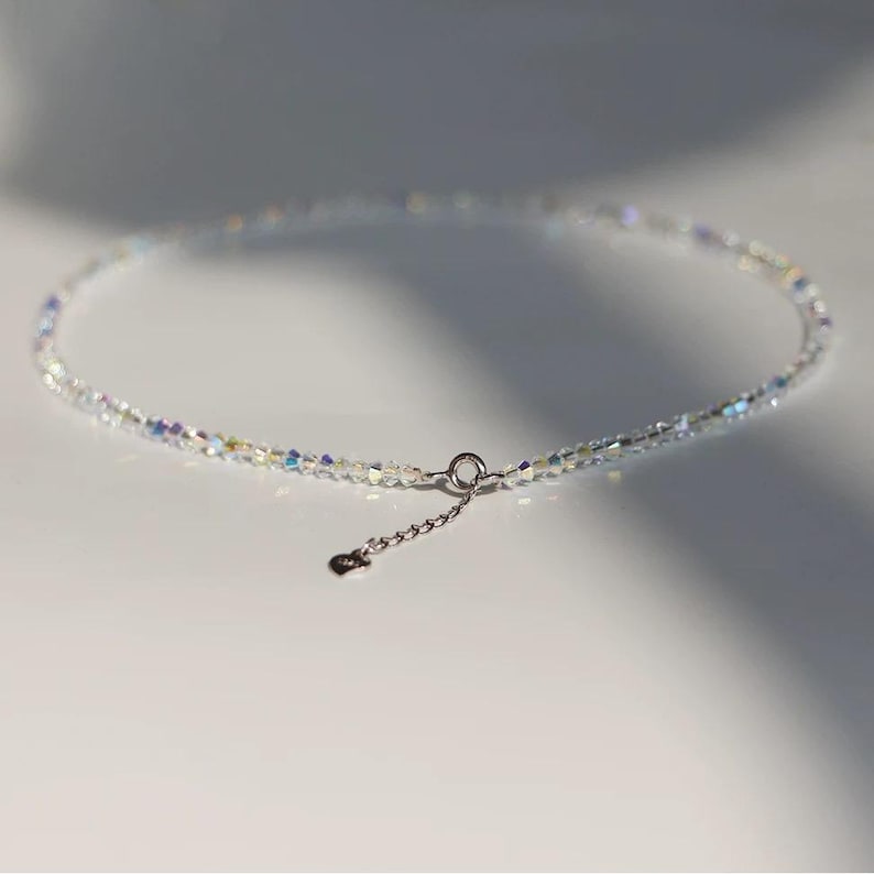 May include: Delicate necklace featuring iridescent, clear crystal beads. The necklace has a silver-toned clasp and a small chain extension with a heart-shaped charm. The beads catch the light, creating a shimmering effect. A minimalist jewelry piece.