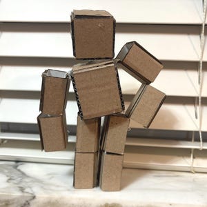 May include: A handmade cardboard figure assembled from small, brown cardboard boxes. The figure has a blocky, geometric design with a head, torso, arms, and legs. The figure is positioned in front of a window with white blinds.