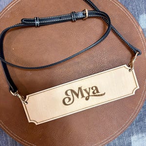 May include: A light-colored wooden sign with the name "Mya" engraved in a decorative font. The sign is suspended by a black leather strap with white stitching and gold-colored hardware. The sign is resting on a brown leather surface.