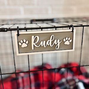May include: A wooden sign for a pet crate, featuring the name "Rudy" in cursive script. The sign has a gray background with paw prints and a white border. It is attached to a black wire crate.