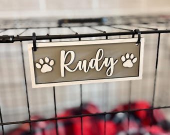 May include: A wooden sign for a pet crate, featuring the name "Rudy" in cursive script. The sign has a gray background with paw prints and a white border. It is attached to a black wire crate.