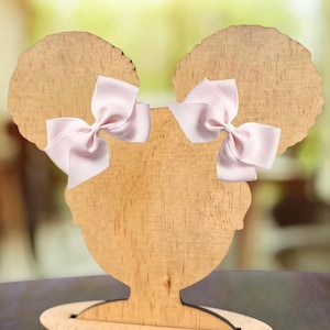 Puede incluir: Expositor de lazos para el pelo de madera con diseño de silueta. El expositor presenta dos lazos de satén rosa claro sujetos a la forma de la cabeza de madera. El expositor es de madera clara y tiene una base redonda.