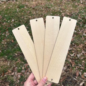 May include: A hand holds four unfinished wooden bookmarks. Each bookmark is rectangular with rounded corners and a small hole at the top for a tassel or string. The wood has a light, natural color.