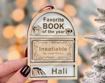 May include: Wooden Christmas ornament with the text "Favorite BOOK of the year" and the book title "Insatiable" by Leigh Rivers. The ornament has a red ribbon for hanging and includes the name "Hali".