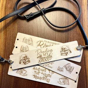 Puede incluir: Marcadores de libros de madera con diseños grabados con temática de libros y texto. Un marcador dice "I'd Rather...", otro dice "Reading my anxiety away", y un tercero dice "Stayed Up All Night Reading". También se ve una correa de cuero negro con cierres metálicos.