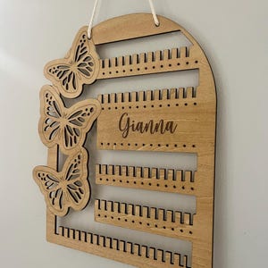 May include: Wooden earring holder with a butterfly design. The light brown holder features three butterfly cutouts on the left. The name "Gianna" is engraved on the front. Multiple rows of holes are designed to hold earrings.