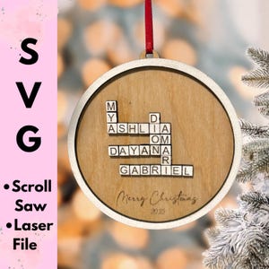 Op de afbeelding: Houten kerstornament met een lichtbruin midden en een crèmekleurige rand. Het ornament heeft namen uitgespeld in Scrabble-achtige tegels. De tekst "Merry Christmas 2025" is eronder gegraveerd. Een rood lint is bevestigd om op te hangen.