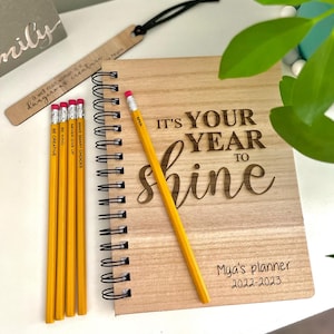 以下が含まれることがあります： 「IT'S YOUR YEAR TO shine」と「Mya's planner 2022-2023」の文字が入った木製プランナー。 黄色の鉛筆、木製のブックマーク、木製の看板が含まれています。 鉛筆にはやる気を起こさせるフレーズが書かれています。