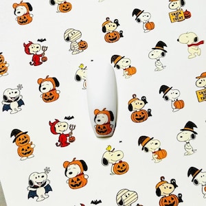 May include: Halloween nail art stickers with Snoopy characters in costumes such as a pumpkin, devil, and mummy. The stickers are on a white background, with a single nail art sample showing a Snoopy pumpkin design.
