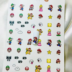 Può includere: Foglio di adesivi per nail art con personaggi di Super Mario, tra cui Mario, Luigi, Principessa Peach e Bowser. Il design incorpora stelle, blocchi punto interrogativo e altri elementi del gioco. Il testo "BEARS CUSTOM CLAW'S" è in alto.