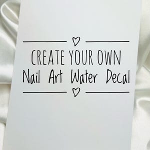 Op de afbeelding: Een rechthoekige witte kaart met de zwart gedrukte tekst CREATE YOUR OWN Nail Art Water Decal, gecentreerd tussen twee horizontale lijnen, elk geaccentueerd met een klein handgetekend hartsymbool boven en onder de hoofdtekst.
