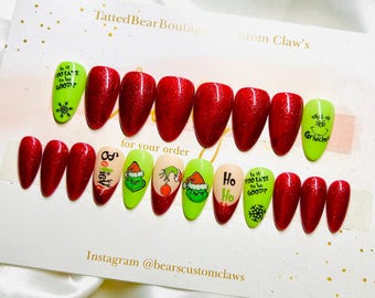 May include: A set of almond-shaped press-on nails featuring a festive theme with glittery red, lime green, and nude colors. Designs include the Grinch character, the text "Is it TOO LATE to be GOOD?", "Ho Ho Ho", "Believe", and "why it up Grinches".