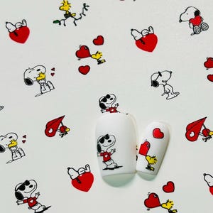 Pu&ograve; includere: Adesivi per nail art con i personaggi di Snoopy e Woodstock con cuori rossi e note musicali. Gli adesivi sono su sfondo bianco, con due unghie dipinte che mostrano i disegni. Il testo "BEARS CUSTOM CLAW'S" &egrave; visibile.