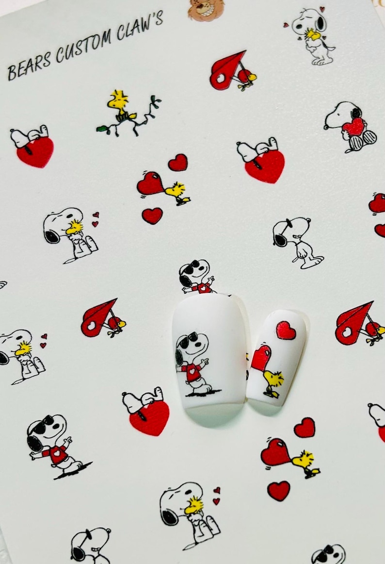 Pu&ograve; includere: Adesivi per nail art con i personaggi di Snoopy e Woodstock con cuori rossi e note musicali. Gli adesivi sono su sfondo bianco, con due unghie dipinte che mostrano i disegni. Il testo "BEARS CUSTOM CLAW'S" &egrave; visibile.
