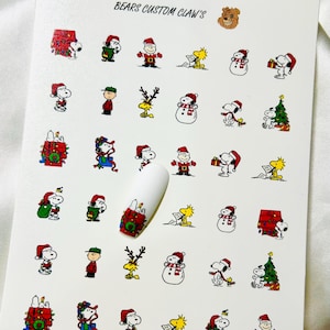 Könnte beinhalten: Bogen mit Nagelkunst-Aufklebern mit Snoopy und seinen Freunden in Weihnachtskleidung. Der Bogen enthält verschiedene Bilder von Snoopy, Woodstock und anderen Charakteren sowie Weihnachtsbäumen und anderen Weihnachtsdekorationen. Der Text "BEARS CUSTOM CLAW'S" ist oben aufgedruckt.