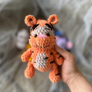 以下が含まれることがあります： 黒い縞模様、グレーと白の腹、ピンクの鼻が付いた、手作りのオレンジ色の編みぐるみトラのおもちゃ。 ぬいぐるみには小さくて丸い耳が付いています。 おもちゃは人の手に持たれています。