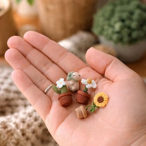 Może przedstawiać: Dłoń trzyma kilka miniaturowych roślin na szydełku, w tym kaktus z białymi kwiatami, małego psa i słonecznik. Rośliny są w brązowych doniczkach. Tło jest rozmyte, z widocznymi innymi roślinami.
