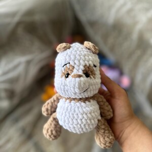 以下が含まれることがあります： 手作りの、ぬいぐるみのようなかぎ針編みの犬のおもちゃ。おもちゃは、耳、鼻、足にタンのアクセントが付いた白い体を持っています。子犬は黒い目をし、縫い付けられた笑顔をしています。おもちゃは人の手で持たれています。