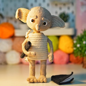 Può includere: Una figura all'uncinetto di un elfo domestico con grandi orecchie, grandi occhi e un lungo naso. La figura è beige con un oggetto nero in mano e un oggetto nero sulla superficie. La figura è in piedi su una superficie bianca.