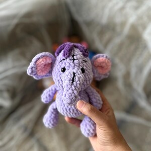 以下が含まれることがあります： ピンク色の内耳と紫色のヘッドピースが付いた、小さなラベンダー色の手編みの象。 手作りのぬいぐるみは黒い目を持ち、手で持たれています。 象は柔らかいぬいぐるみです。