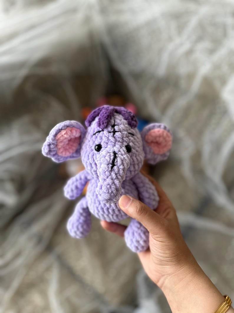 以下が含まれることがあります： ピンク色の内耳と紫色のヘッドピースが付いた、小さなラベンダー色の手編みの象。 手作りのぬいぐるみは黒い目を持ち、手で持たれています。 象は柔らかいぬいぐるみです。