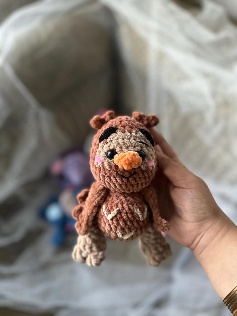 以下が含まれることがあります： 茶色、ベージュ、オレンジ色の手作りの編みぐるみフクロウ人形。 フクロウは黒い目、オレンジ色のくちばし、ピンクの頬をしています。 人形は質感のある外観をしています。
