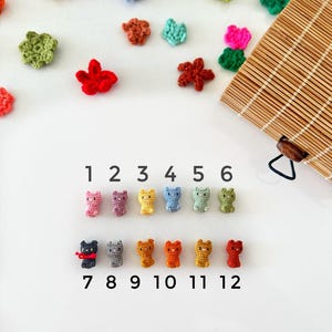 Pode incluir: Doze figurinhas de gato de crochê em várias cores, numeradas de 1 a 12. Os gatos estão dispostos em duas filas, com a primeira fila com cores pastel e a segunda fila com cores mais vibrantes.