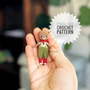 Può includere: Un orso di crochet marrone che indossa una sciarpa rossa e una salopette verde. L'orso è tenuto in una mano. L'immagine include il testo "ENGLISH CROCHET PATTERN".