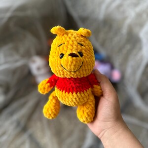 以下が含まれることがあります： 手作りの黄色と赤のかぎ針編みのくまのプーさんのぬいぐるみ。ぬいぐるみは笑顔の顔、小さな黒い目、赤いシャツをしています。おもちゃは手で持たれています。