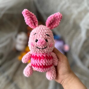 以下が含まれることがあります： 赤とピンクのストライプのシャツを着た、手作りのピンクのかぎ針編みの豚のぬいぐるみ。豚は大きなピンクの耳、小さな鼻、黒い刺繍の目を持っています。ぬいぐるみは手で持たれています。