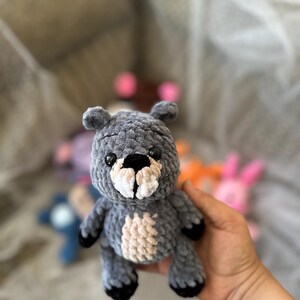 以下が含まれることがあります： 黒い足と黒い鼻が付いた、手作りの灰色の編み物ベア。 クリーミーな色の胸と小さな黒い目があります。 他のカラフルな編み物動物が背景に見えます。