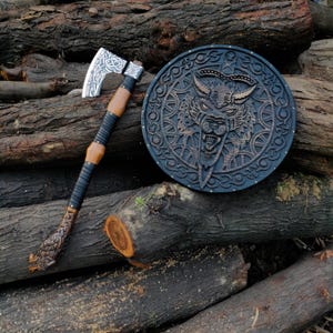 May include: A Viking-style axe with a silver blade and wooden handle, next to a round wooden shield with a carved wolf design. The axe features intricate knotwork on the blade and handle. The shield has a dark finish.