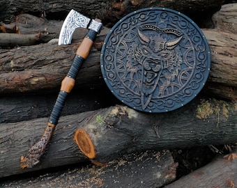 May include: A Viking-style axe with a silver blade and wooden handle, next to a round wooden shield with a carved wolf design. The axe features intricate knotwork on the blade and handle. The shield has a dark finish.