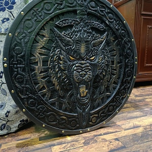 Peut inclure: Un bouclier rond noir avec un motif détaillé de tête de loup au centre. Le loup a des yeux jaunes brillants et une gueule ouverte. Le bouclier est orné d'entrelacs celtiques complexes et de clous couleur laiton sur le bord.