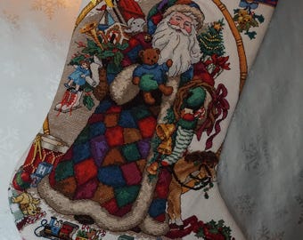 Könnte beinhalten: Ein cremefarbener Weihnachtsstrumpf mit einem detaillierten Kreuzstichmotiv des Weihnachtsmanns, der einen Teddybären hält. Der Strumpf zeigt einen bunten Patchwork-Mantel, verschiedene Spielzeuge, einen Zug und eine Holztruhe mit gelber Schlaufe.