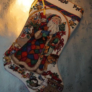 May include: Embroidered Christmas stocking featuring Santa Claus holding toys and a teddy bear. The stocking has a colorful patchwork design on Santa's coat and is trimmed with a gold ribbon. The background is a light blue with snowflake patterns.