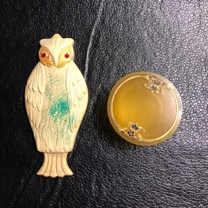 May include: A white ceramic owl figurine with red eyes and a green and white design. The owl is standing on a black surface next to a round, yellow, ceramic trinket box with gold accents and a floral design.