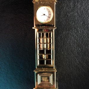 May include: A gold-toned miniature grandfather clock with a white face and black hands. The clock has a decorative top and a glass front.