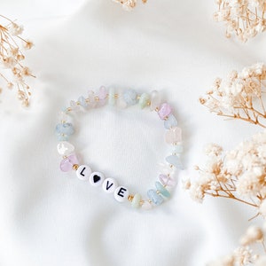 May include: A bracelet made of light blue, pink and green stones with gold accents. The bracelet has the word "LOVE" spelled out with white beads and a black heart.
